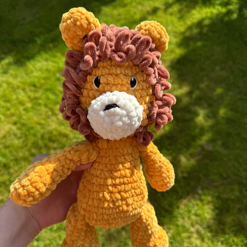Un lion en peluche jaune et brun fait au crochet, avec une crinière et des oreilles, tenu dans une main contre un fond de gazon.